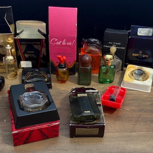 May include: A collection of various miniature perfume bottles and their corresponding boxes, including brands like Moschino, Christian Lacroix, Boucheron, and Quorum. The assortment features glass bottles in diverse shapes, colors, and sizes.