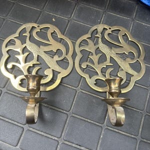 May include: Two brass peacock candle holders. Each holder features a peacock design within an ornate, openwork frame. The candle holders have a small cup to hold the candle and a loop for hanging. The brass has a warm, aged patina.