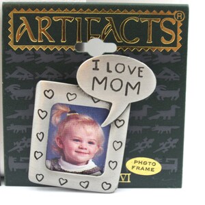 May include: A silver photo frame with a heart border and a speech bubble that says "I love Mom". The frame is on a green background with the word "ARTIFACTS" in gold letters. The frame is labeled "PHOTO FRAME".