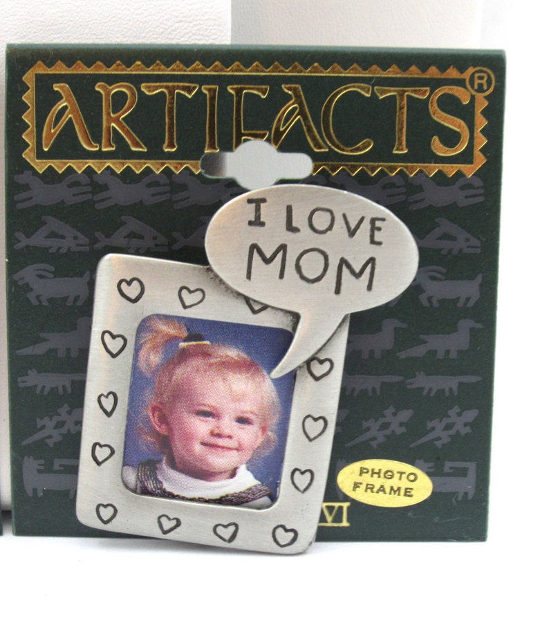 May include: A silver photo frame with a heart border and a speech bubble that says "I love Mom". The frame is on a green background with the word "ARTIFACTS" in gold letters. The frame is labeled "PHOTO FRAME".
