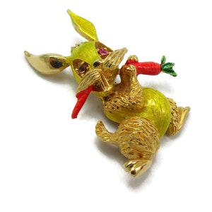 Puede incluir: Una broche de conejo de esmalte amarillo con detalles dorados, que lleva una zanahoria roja y verde. El conejo tiene un ojo de pedrería.