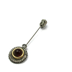 May include: A silver pin with a round, dark red gemstone surrounded by a gold band and a silver rope design. The pin has a clear rhinestone on the stem.