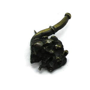 Könnte beinhalten: Eine bronzene Anstecknadel in Form eines kleinen Tieres mit einer langen, gebogenen Pfeife. Das Tier sitzt auf seinen Hinterbeinen und hält die Pfeife im Maul. Die Anstecknadel ist im Vintage-Stil gehalten und wäre eine großartige Ergänzung für jede Sammlung.