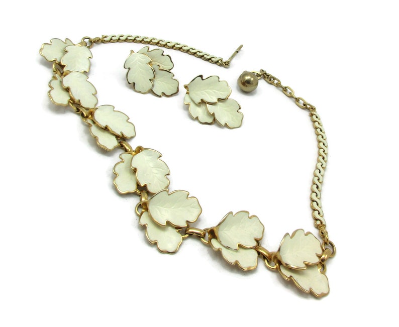 May include: A gold chain necklace with white enamel leaf charms and matching earrings. The necklace has a lobster clasp closure.