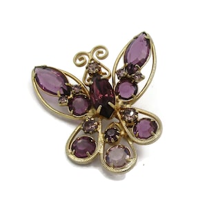 Puede incluir: Un broche de mariposa de tono dorado con diamantes de imitación morados. La mariposa tiene un diseño detallado con alas intrincadas y un cuerpo delicado.