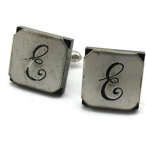 Puede incluir: Un par de gemelos de plata con detalles en esmalte negro. Cada gemelo presenta una letra "E" estilizada en negro.
