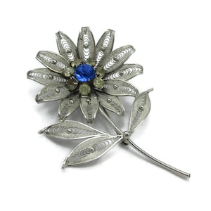 Könnte beinhalten: Eine silberne Filigran-Blumenbrosche mit einem blauen Strassstein in der Mitte und kleineren klaren Strasssteinen. Die Blume hat einen Stiel und Blätter.