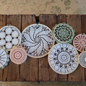 May include: A collection of eight crocheted doilies in various sizes and colors, including white, green, and pink, are displayed on a wooden surface. The doilies are circular and feature intricate patterns.