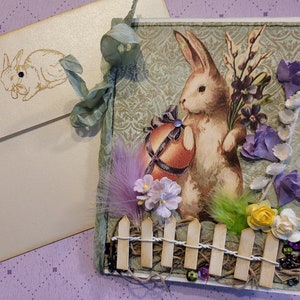 Carte de lapin de Pâques dans le jardin avec clôture en bois, fleurs de chapellerie vintage et plumes, couleurs printanières