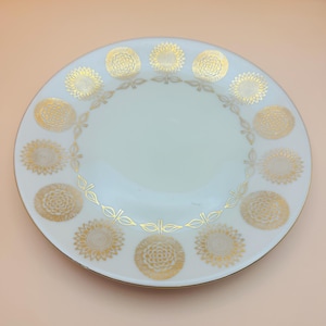 May include: White ceramic plate with a gold floral design. The plate features a ring of gold sunflowers around the edge and a gold trim. The plate has a smooth, glossy finish and is set against a light peach background.