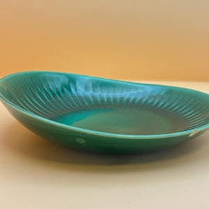 Könnte beinhalten: Eine grüne Keramik-Servierschale mit gerilltem Design. Die ovale Schale hat eine glänzende Oberfläche und eine tiefgrüne Farbe. Die Schale ist leer und steht auf einer hellorangen Oberfläche.