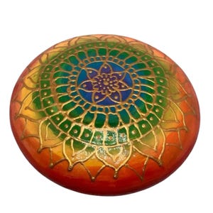 Op de afbeelding: Een ronde, beschilderde steen met een regenboogmandala-ontwerp. De mandala heeft een centrale bloem met gouden accenten en een buitenste ring in groen, blauw, paars en geel.