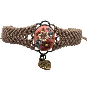 Op de afbeelding: Handgemaakte macramé armband met een centraal ovaalvormig roze bloemmotief. Het ontwerp heeft kleine, kleurrijke bloemen en een bronzen metalen frame. Een kleine hartvormige bedel met de tekst "made with love" hangt eronder.