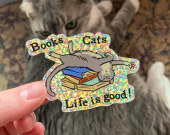 Könnte beinhalten: Ein glitzernder Aufkleber mit einer Katze, die auf einem Stapel Bücher liegt. Der Aufkleber lautet "Books & Cats Life is good!"