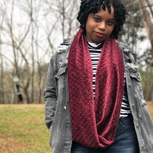 May include: A burgundy infinity scarf with a textured, dotted pattern. The scarf is draped around the neck, showcasing its soft, plush material. The person is wearing a denim jacket and a black and white striped shirt.