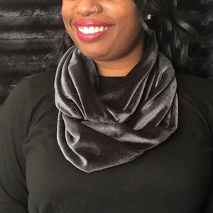 May include: A gray velvet infinity scarf. The scarf is worn by a person with dark hair and a black shirt.