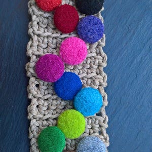 Peut inclure: Gros plan sur une variété de boules de feutre colorées disposées en rangée sur un fond de jute tissé. Les boules sont dans des tons de rouge, orange, jaune, vert, bleu, violet, rose et marron.