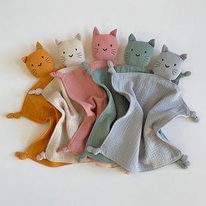 May include: Five plush cat toys with attached blankets. The cats are in various colors: orange, beige, pink, green, and gray. Each cat has a simple black embroidered face.