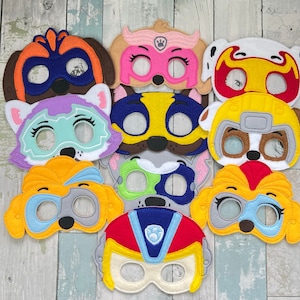 May include: A selection of colourful felt masks depicting cartoon dog characters. The masks are in shades of brown, pink, yellow, blue, and grey, with eye holes and character-specific details. The masks are designed for children's play or costume use.