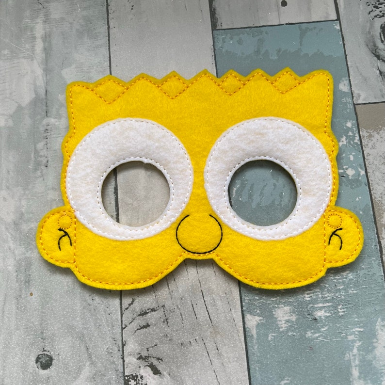 Puede incluir: Una m&aacute;scara de fieltro amarilla con ojos blancos y una sonrisa negra. La m&aacute;scara est&aacute; dise&ntilde;ada para parecerse a un personaje de dibujos animados con pelo amarillo puntiagudo.