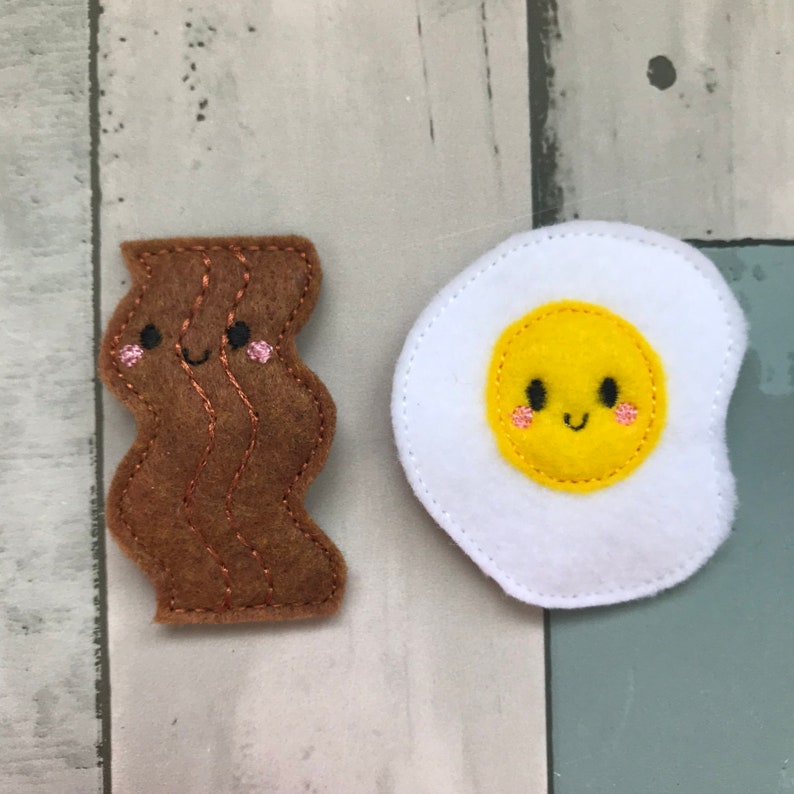 May include: Two felt food items, a brown bacon and a white egg with a yellow yolk, both with embroidered smiling faces.