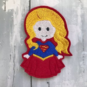 May include: Embroidered felt patch of a cartoon superhero with blonde hair, wearing a red skirt, blue shirt with a yellow Superman logo, and a yellow belt.