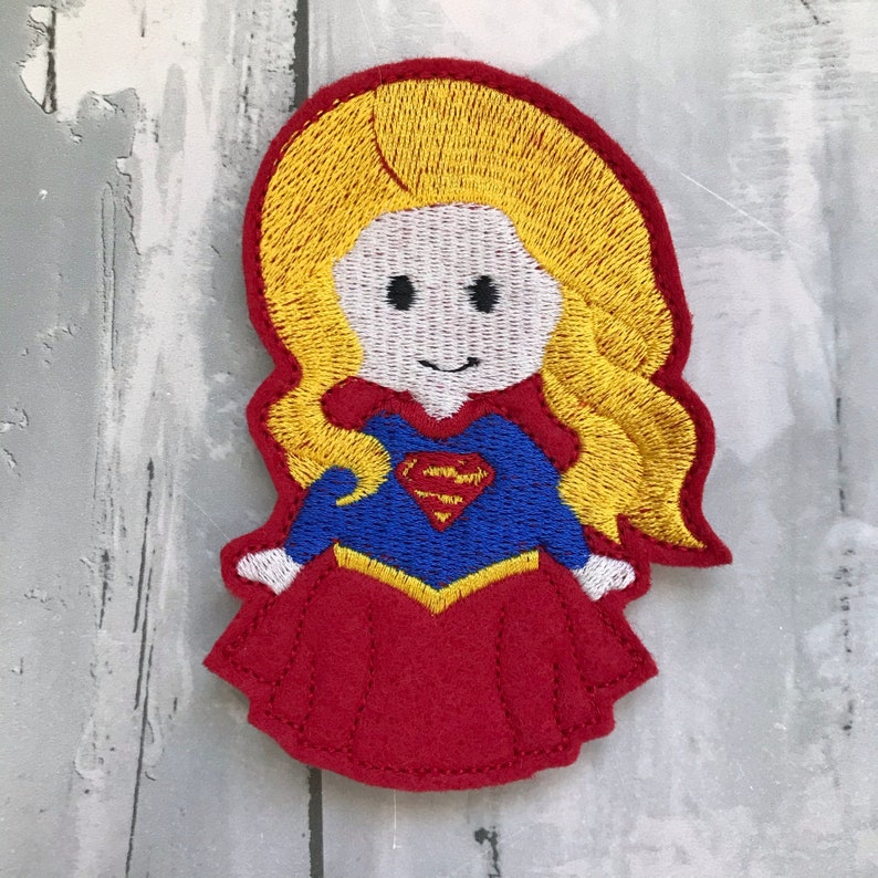 May include: Embroidered felt patch of a cartoon superhero with blonde hair, wearing a red skirt, blue shirt with a yellow Superman logo, and a yellow belt.