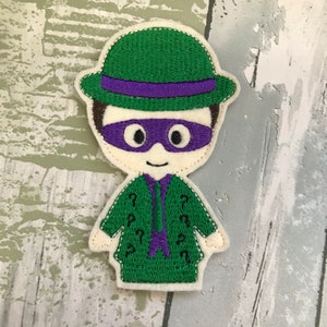May include: Green felt applique of the Riddler from Batman, wearing a green hat with a purple band, a purple mask, and a green suit with question marks.