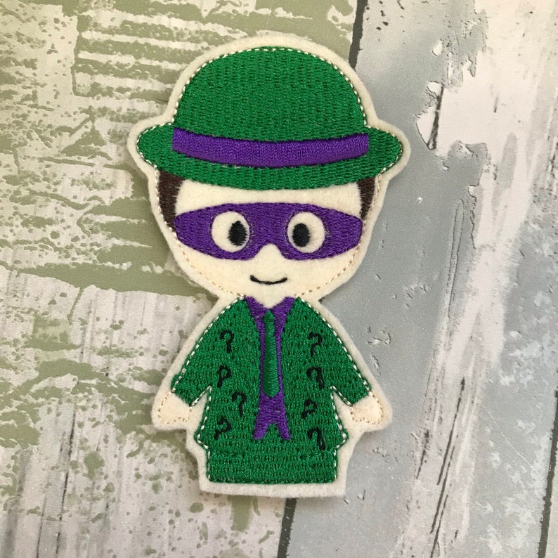 May include: Green felt applique of the Riddler from Batman, wearing a green hat with a purple band, a purple mask, and a green suit with question marks.