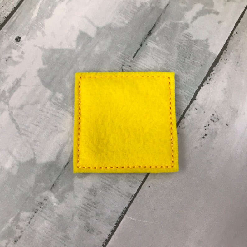 Puede incluir: Un peque&ntilde;o cuadrado de fieltro amarillo brillante con un borde cosido.