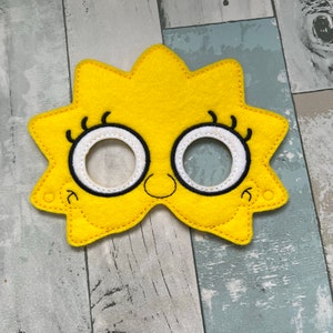 Puede incluir: M&aacute;scara de fieltro amarilla con ojos y pesta&ntilde;as negros, una sonrisa blanca y dos peque&ntilde;os c&iacute;rculos a los lados. La m&aacute;scara tiene forma de estrella.