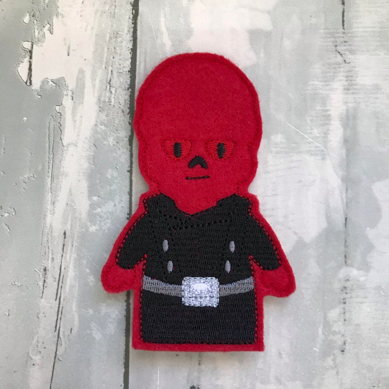 May include: Red felt finger puppet of a character with a bald head, black eyes, and a black outfit. The character is wearing a black belt with a silver buckle.