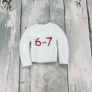 May include: A small, white, fluffy jumper with a crew neck and long sleeves. The number "6-7" is embroidered in red on the front. The jumper is displayed on a light grey and white wooden surface.