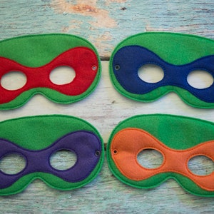 May include: Four felt superhero masks with green outer edges and different coloured inner sections. The masks have two eye holes and are sewn with a decorative stitch.