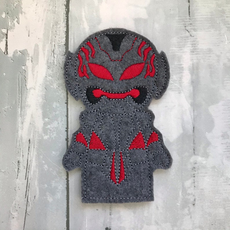 May include: A gray felt finger puppet of a superhero with red eyes and a red design on the chest.