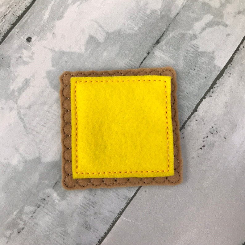 Puede incluir: Un juguete de fieltro con forma de galleta cuadrada con un cuadrado de fieltro amarillo en el interior. La galleta es marr&oacute;n y tiene un borde cosido.