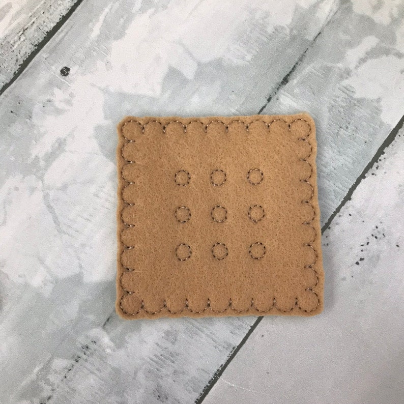 Puede incluir: Un cuadrado de fieltro marr&oacute;n con un borde festoneado y nueve c&iacute;rculos cosidos en una cuadr&iacute;cula de 3x3.