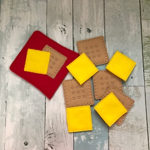 Puede incluir: Un conjunto de piezas de juego de comida de fieltro, que incluyen queso amarillo y galletas marrones. Las piezas est&aacute;n dispuestas sobre un fondo de fieltro rojo.
