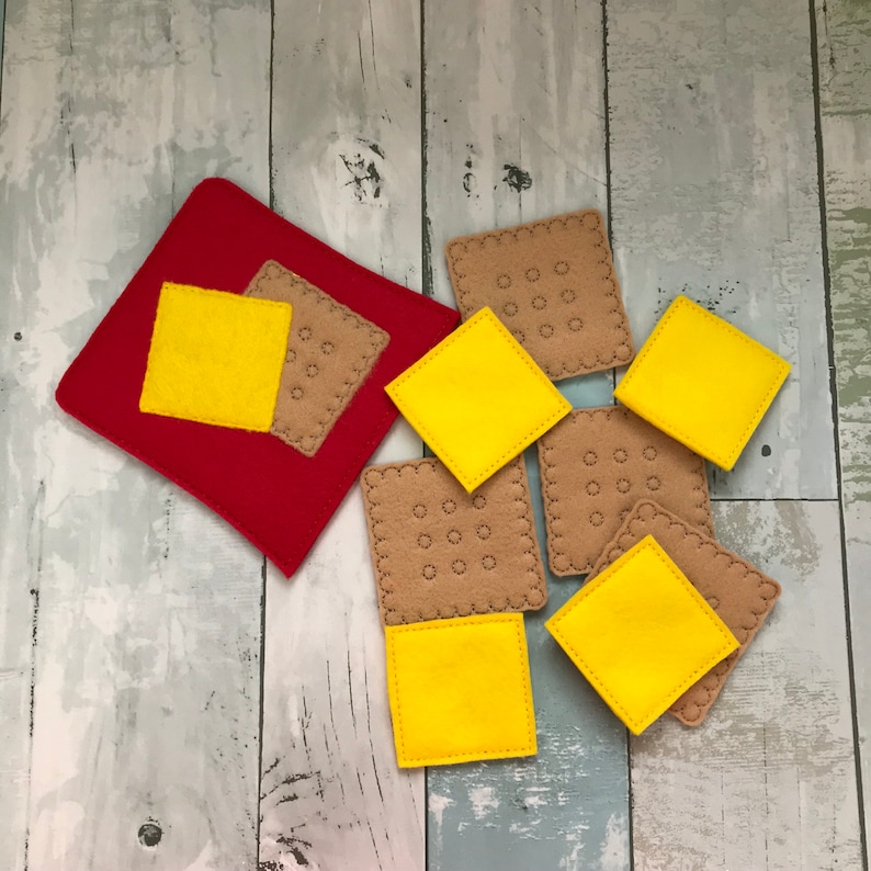 Puede incluir: Un conjunto de piezas de juego de comida de fieltro, que incluyen queso amarillo y galletas marrones. Las piezas est&aacute;n dispuestas sobre un fondo de fieltro rojo.