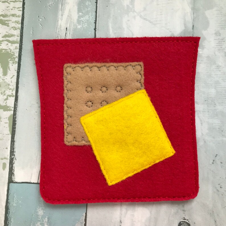 Puede incluir: Un juego de comida de fieltro que incluye un bolsillo de fieltro rojo con una galleta de fieltro marr&oacute;n y una rebanada de queso de fieltro amarillo en su interior.