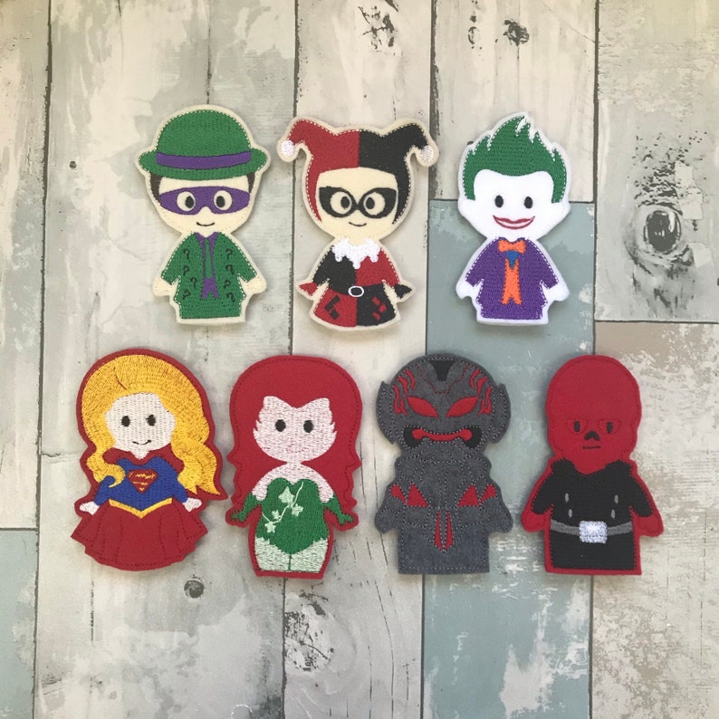 May include: Set of 8 felt finger puppets featuring characters from DC Comics. The puppets are red, green, black, gray, and purple. The characters include Harley Quinn, The Joker, Poison Ivy, The Riddler, Superman, Batman, and Wonder Woman.