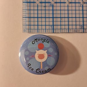 May include: A round button with a light blue background and the words "Moogle Ice Cream" in a whimsical font. The button features a cartoon character with a red pom-pom, white face, and purple wings. A ruler is in the background.