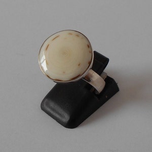 May include: A white and brown ring with a round, textured stone. The ring is on a black ring holder.