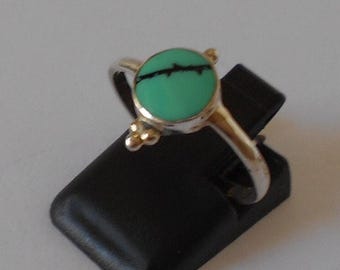 May include: A silver ring with a turquoise stone set in a round bezel. The stone has a black vein running through it. The ring has three small gold beads on the band.