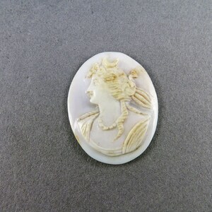 Puede incluir: Colgante de camafeo ovalado con el perfil de una mujer con cabello suelto y un collar. El relieve tallado es en tonos crema y beige, sobre un fondo p&aacute;lido. Una pieza de joyer&iacute;a cl&aacute;sica.