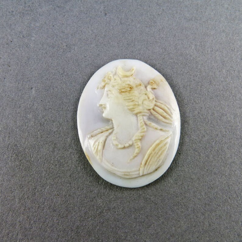 Puede incluir: Colgante de camafeo ovalado con el perfil de una mujer con cabello suelto y un collar. El relieve tallado es en tonos crema y beige, sobre un fondo p&aacute;lido. Una pieza de joyer&iacute;a cl&aacute;sica.