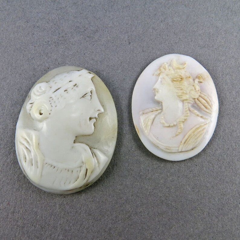 Puede incluir: Dos colgantes de camafeo ovalados. Los camafeos est&aacute;n tallados en relieve con perfiles de mujeres. El camafeo de la izquierda es de color blanco cremoso, mientras que el de la derecha es de un blanco rosado p&aacute;lido. Ambos est&aacute;n sobre un fondo gris.