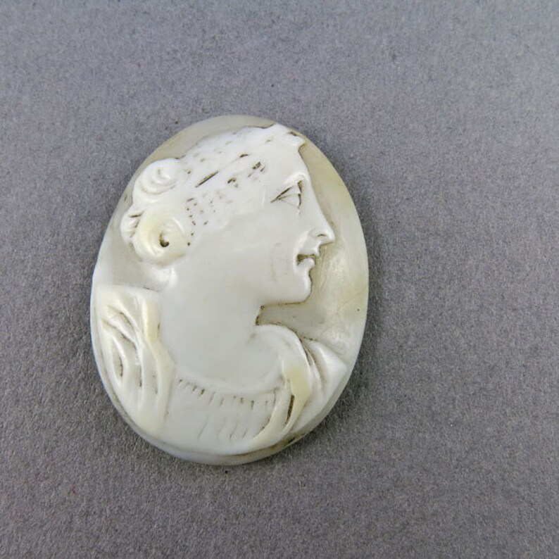 Puede incluir: Colgante de camafeo ovalado que representa el perfil de una mujer. El tallado es de color blanco crema, con el pelo de la mujer recogido. El fondo es de color blanco roto. El colgante es probablemente de concha o material similar.