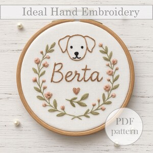 Könnte beinhalten: Ein bestickter Reifen mit einem weißen Hund, dem Namen Berta und einem Blumenkranz in Rosa und Grün. Der Reifen ist aus Holz und hat einen weißen Stoffhintergrund.