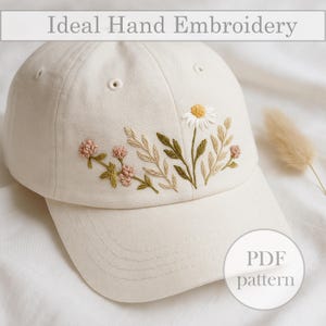Può includere: Cappellino da baseball color crema con ricamo floreale fatto a mano. Il ricamo presenta una margherita bianca, fiori rosa e foglie verdi e beige. Il testo "Ideal Hand Embroidery" è nella parte superiore dell'immagine e un grafico "PDF pattern" è in basso a destra.